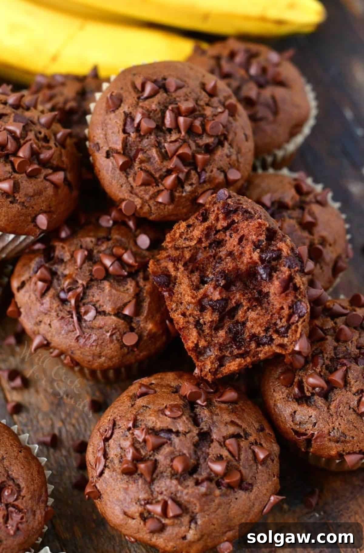 Several rich, moist chocolate banana muffins on a rustic wooden board, with one broken in half to reveal its tender interior and melty chocolate chips.