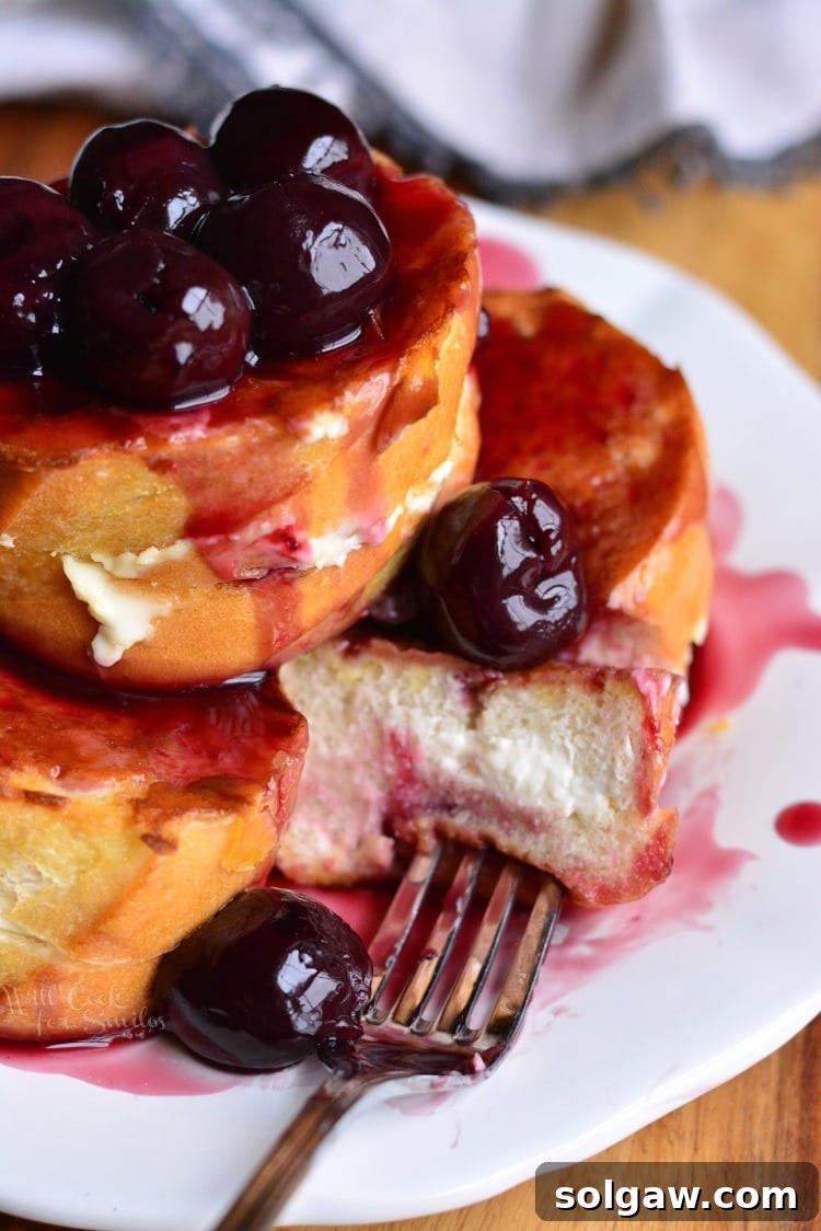 A slice of cherry cheesecake stuffed french toast on a white plate with a fork, showing the creamy filling and cherry topping.