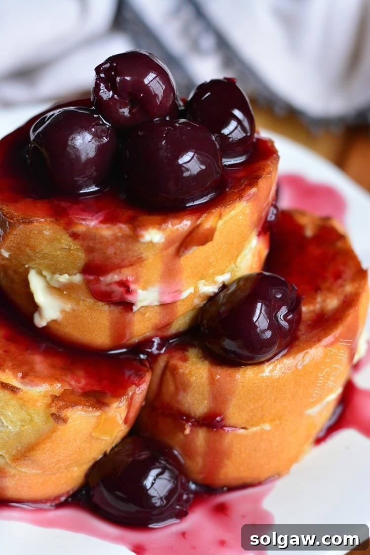 A stack of golden French toast slices, each filled with creamy cheesecake and drizzled with glistening dark cherry sauce, on a white plate.