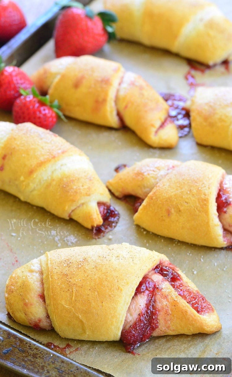 Six golden-brown strawberry vanilla crescent rolls on a baking sheet, surrounded by fresh red strawberries, ready to be served.