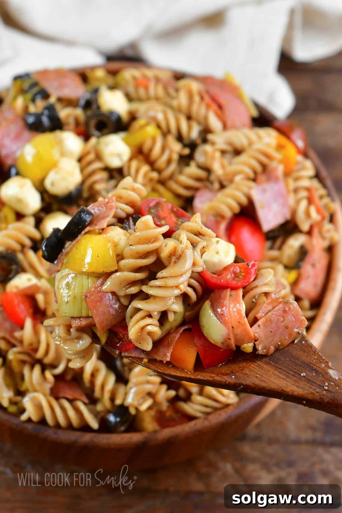 A delightful antipasto pasta salad, featuring rotini, salami, mozzarella balls, black olives, artichoke hearts, pepperoncini, and cherry tomatoes, elegantly presented on a wooden spoon next to a rustic bowl.