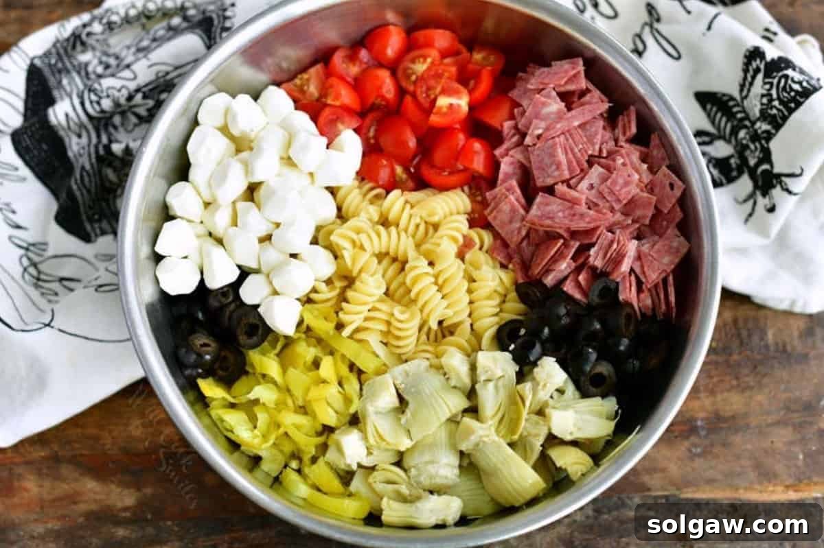 Assorted ingredients for Antipasto Pasta Salad, including cooked pasta, salami, mozzarella, black olives, artichoke hearts, pepperoncini, and cherry tomatoes, gathered in a large metal mixing bowl before being tossed.