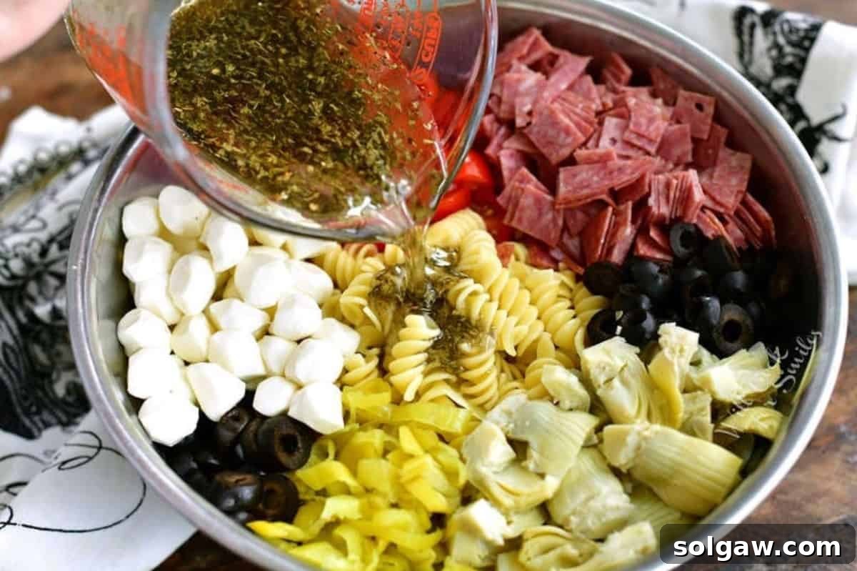 A hand pouring a rich balsamic herb dressing over the colorful ingredients of Antipasto Pasta Salad in a metal mixing bowl, ready to be tossed.