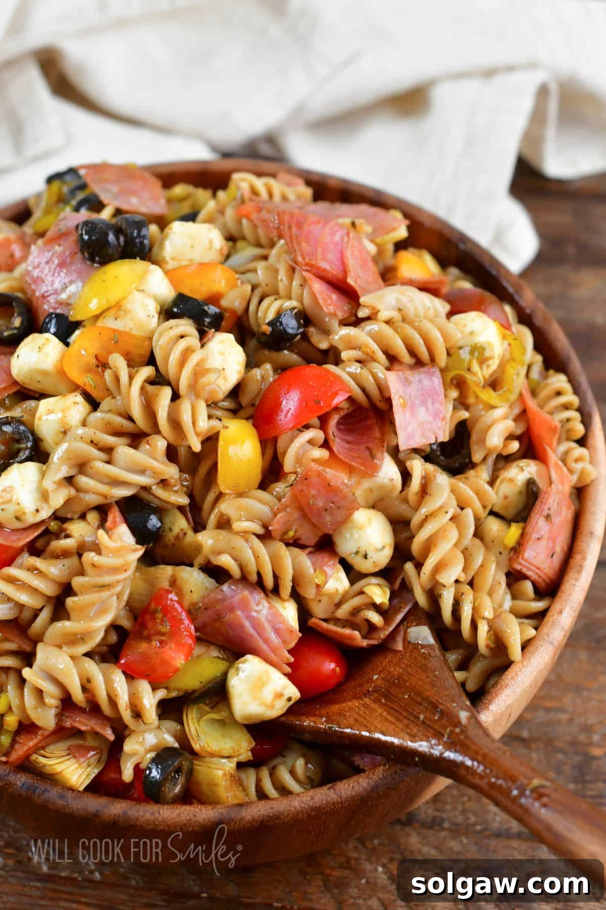 A wooden spoon scooping out a generous portion of Antipasto Pasta Salad from a large wooden bowl, showcasing the medley of pasta, meats, cheeses, and vegetables.