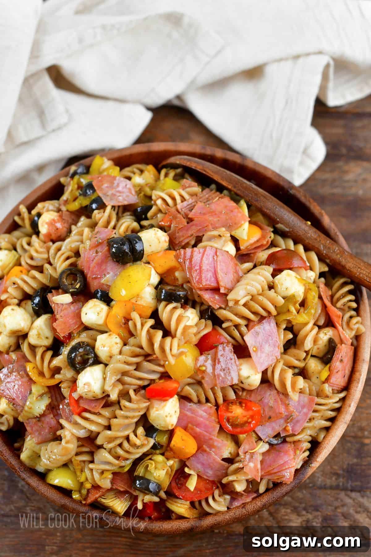 A beautifully presented Italian Antipasto Pasta Salad in a wooden bowl, accompanied by a wooden spoon and a rustic towel, ready to be served.