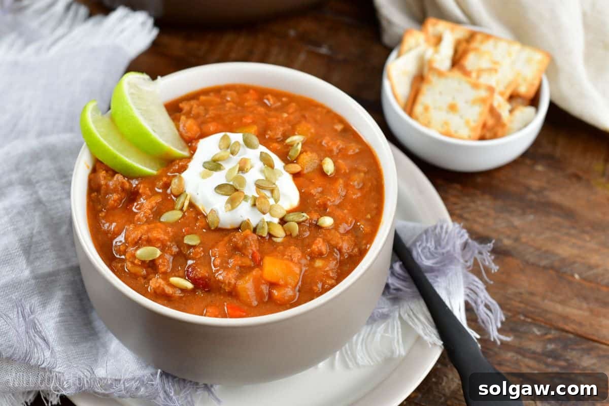 A vibrant bowl of pumpkin chili, artfully garnished with a swirl of sour cream, crunchy pumpkin seeds, and a fresh lime wedge, served alongside a bowl of crackers on a plate.