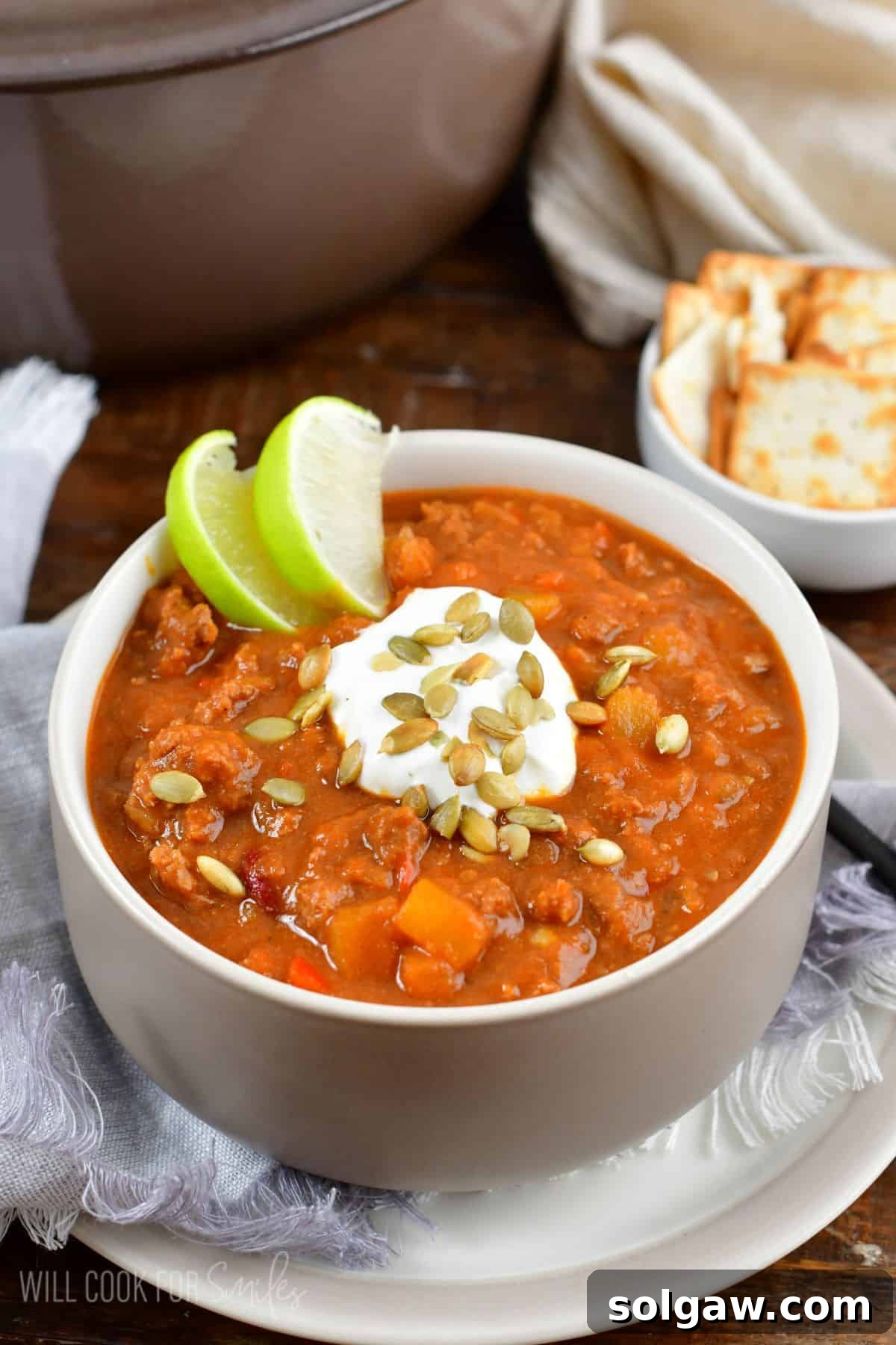 An appetizing bowl of pumpkin chili, garnished with sour cream, pumpkin seeds, and a lime wedge, placed on a white plate.