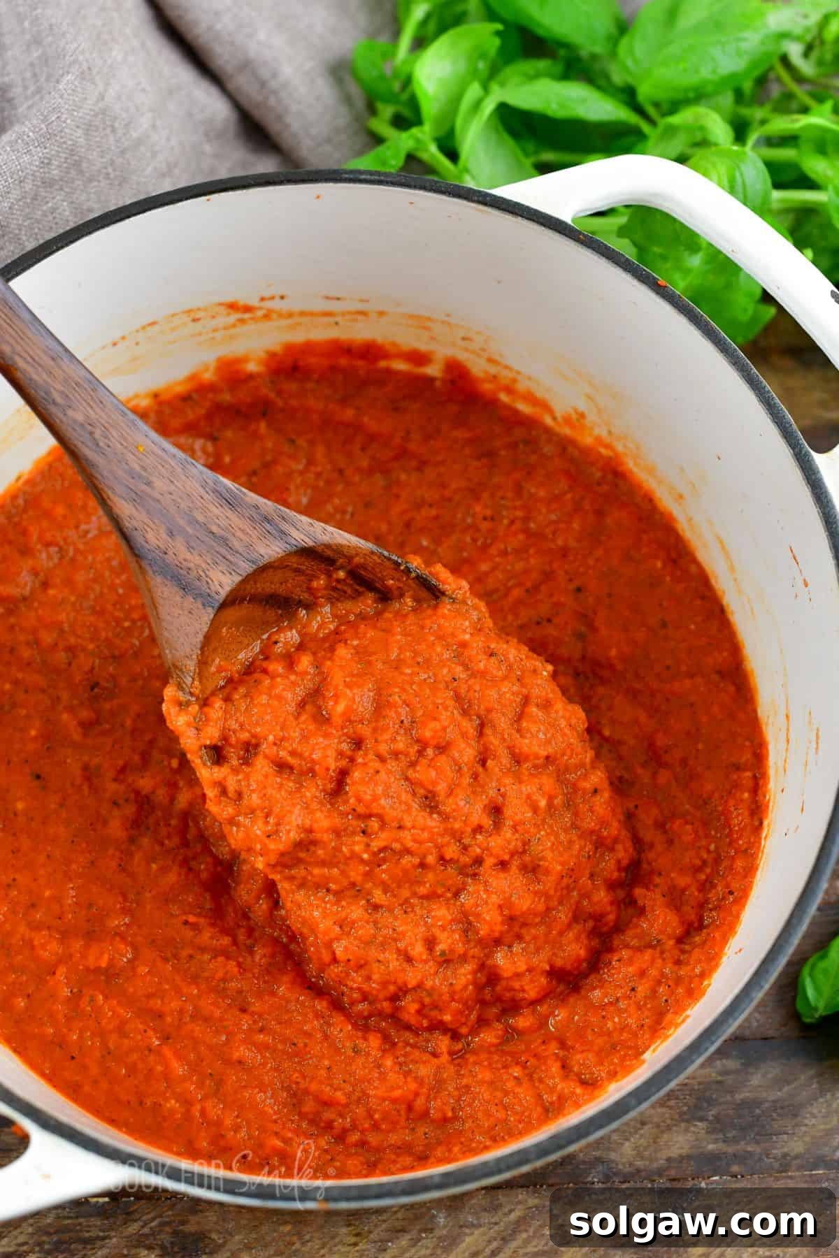 A pot of vibrant homemade spaghetti sauce, richly colored, with a wooden spoon lifting a portion, garnished with fresh basil leaves on a wooden surface.