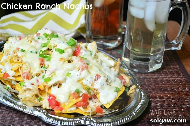 A vibrant platter of Chicken Ranch nachos, freshly baked and garnished with green onions, accompanied by two refreshing drinks.