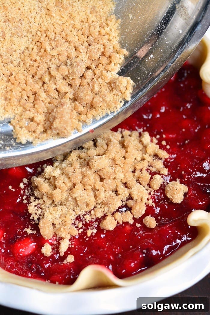 A hand pouring golden streusel topping over the fruit filling of an Apple Cranberry Pie before baking, showing the texture.
