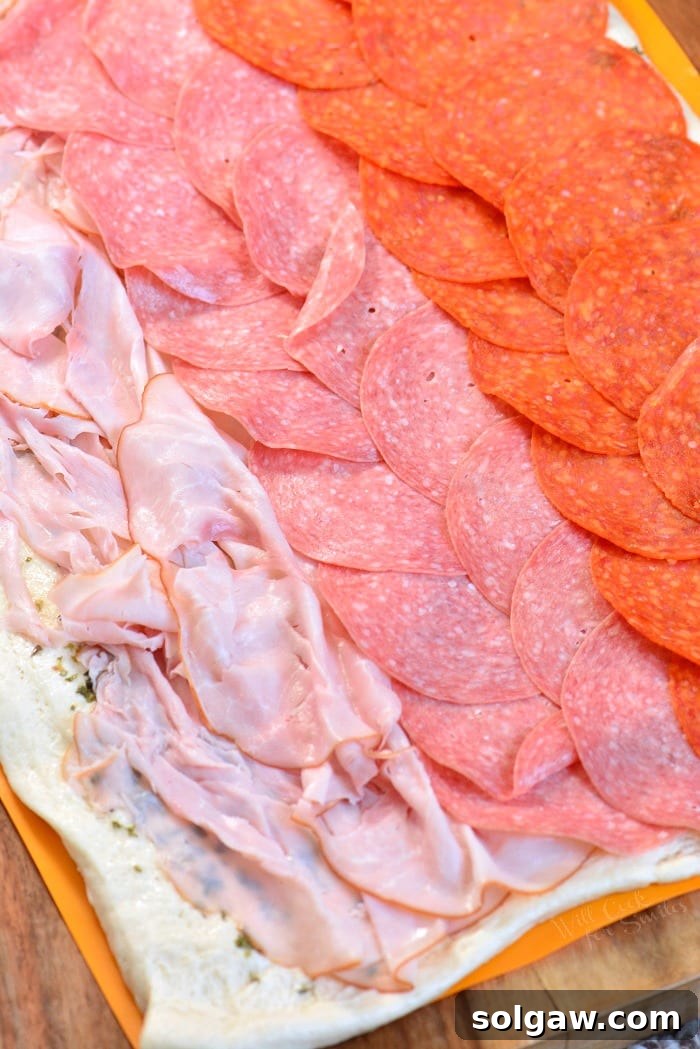 Layering ham, salami, and pepperoni over a stretched pizza dough in preparation for rolling.