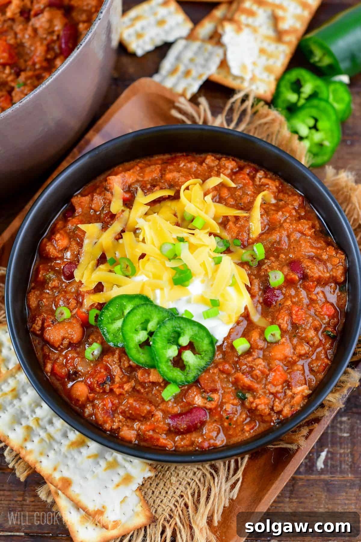 A close-up of a rich, homemade beef chili served in a black bowl, beautifully topped with sour cream, shredded cheese, fresh green onions, and vibrant jalapeños.
