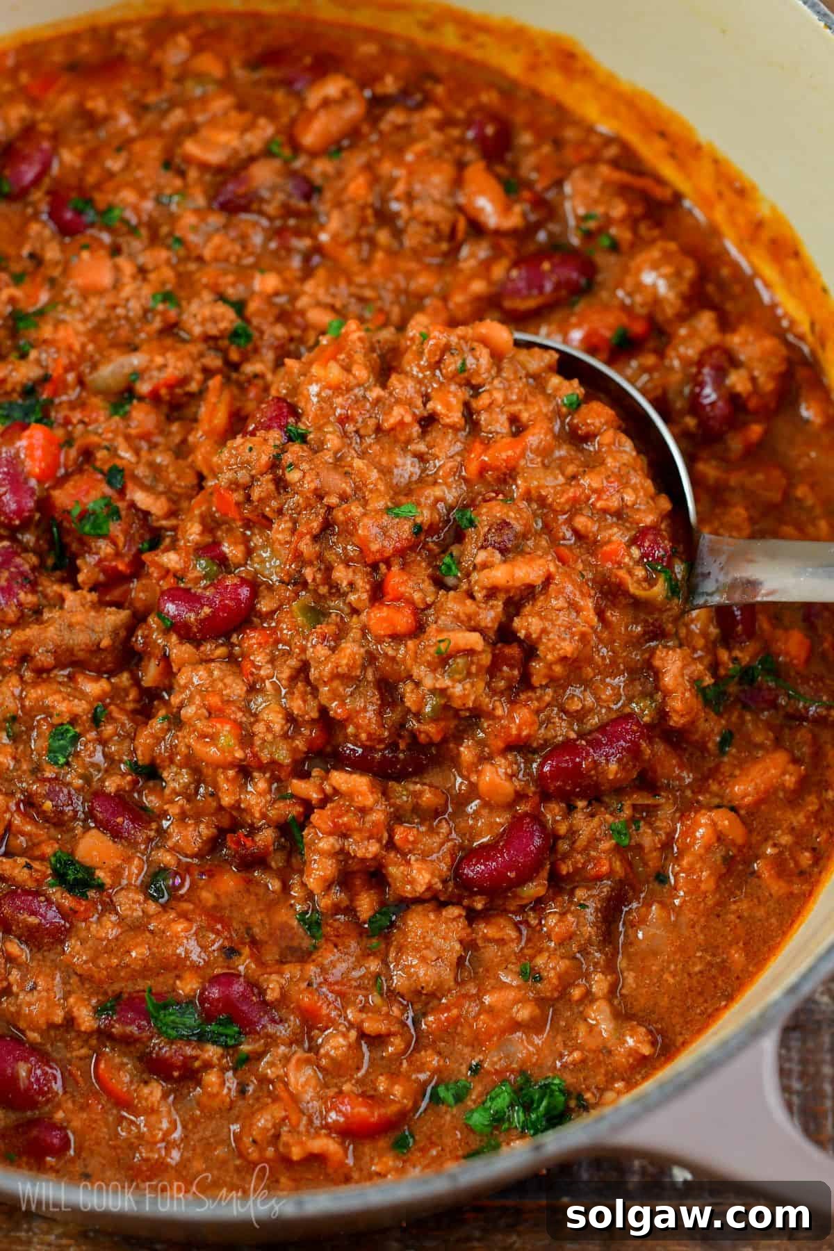 A silver ladle expertly scooping a generous portion of rich, homemade beef chili from a large pot, showcasing its thick consistency and hearty ingredients.