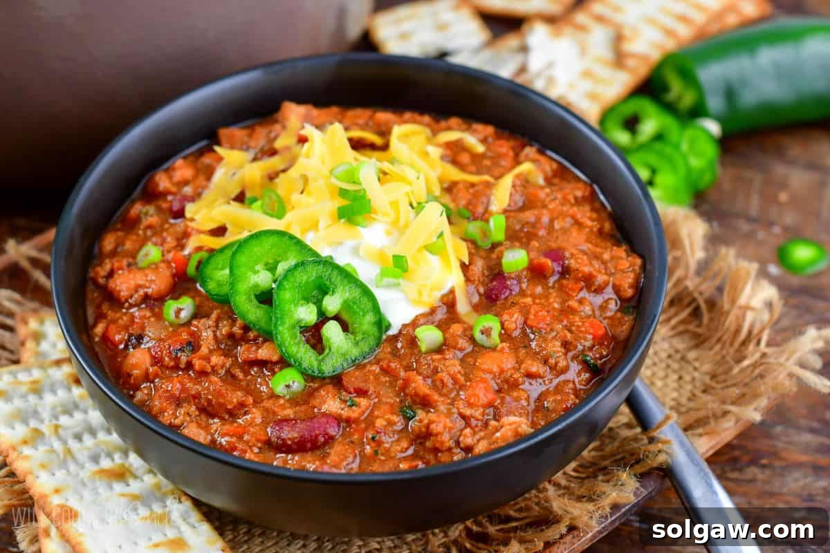 A delicious bowl of beef chili garnished with a generous dollop of sour cream, shredded cheese, fresh green onions, and fiery jalapeños, with a spoon ready for digging in.