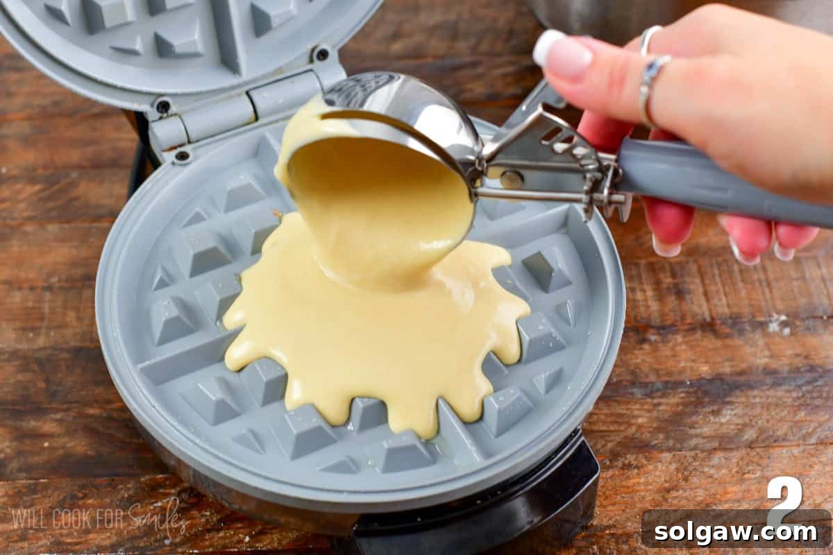 Waffle batter being scooped from a bowl into a preheated waffle maker using an ice cream scoop.