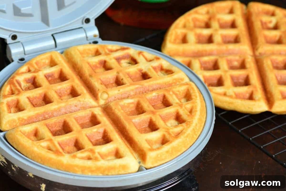 A freshly cooked waffle sits inside a waffle maker, with another perfect waffle resting on a wire rack nearby.