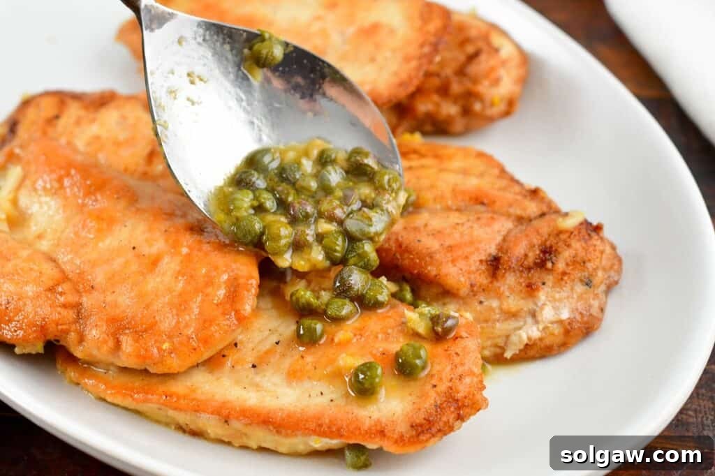 A hand using a spoon to spread the vibrant lemon-butter caper sauce over a portion of seared chicken piccata on a plate.