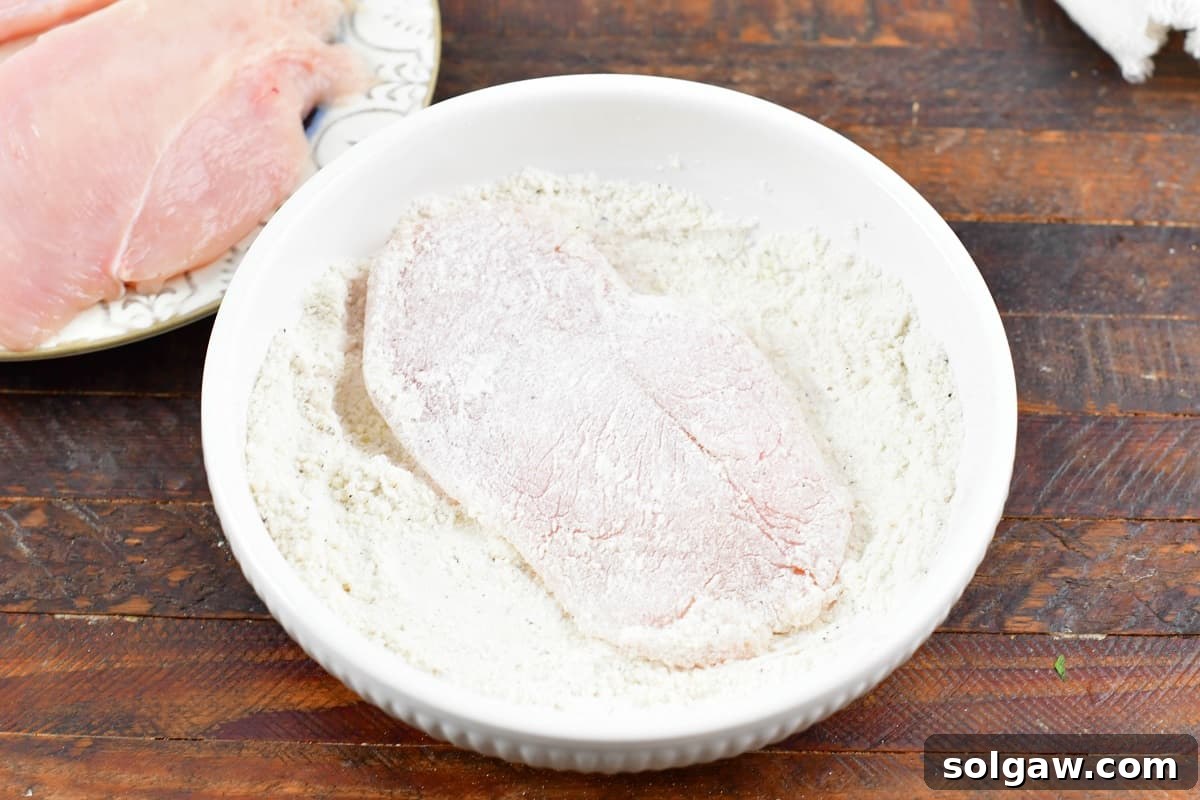 Dredging a chicken cutlet in a bowl of seasoned flour and parmesan cheese mixture, ensuring an even coating.