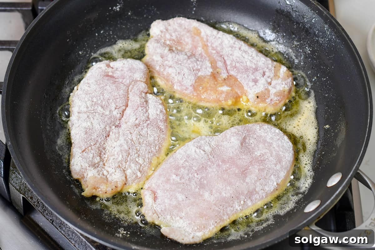 Three dredged chicken cutlets sizzling in a hot pan, developing a beautiful golden-brown sear.