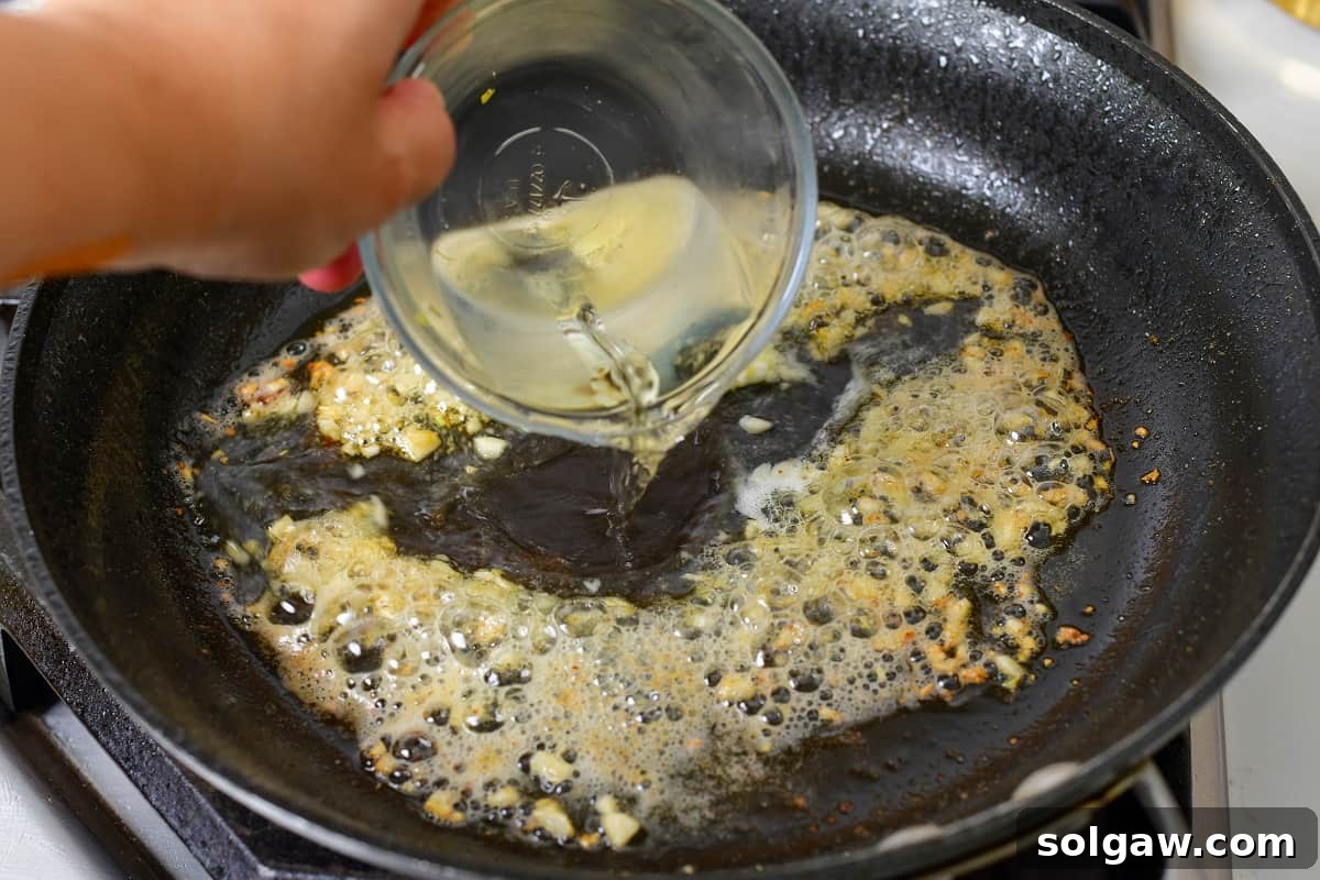 Pouring dry white wine into the hot pan with garlic, beginning the deglazing process for the piccata sauce.
