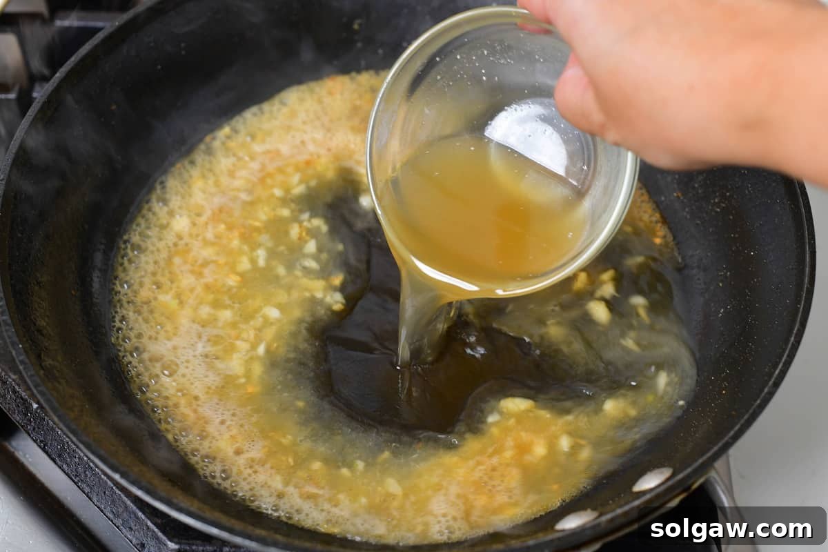 Adding chicken stock to the simmering wine and garlic mixture in the pan, forming the base of the piccata sauce.