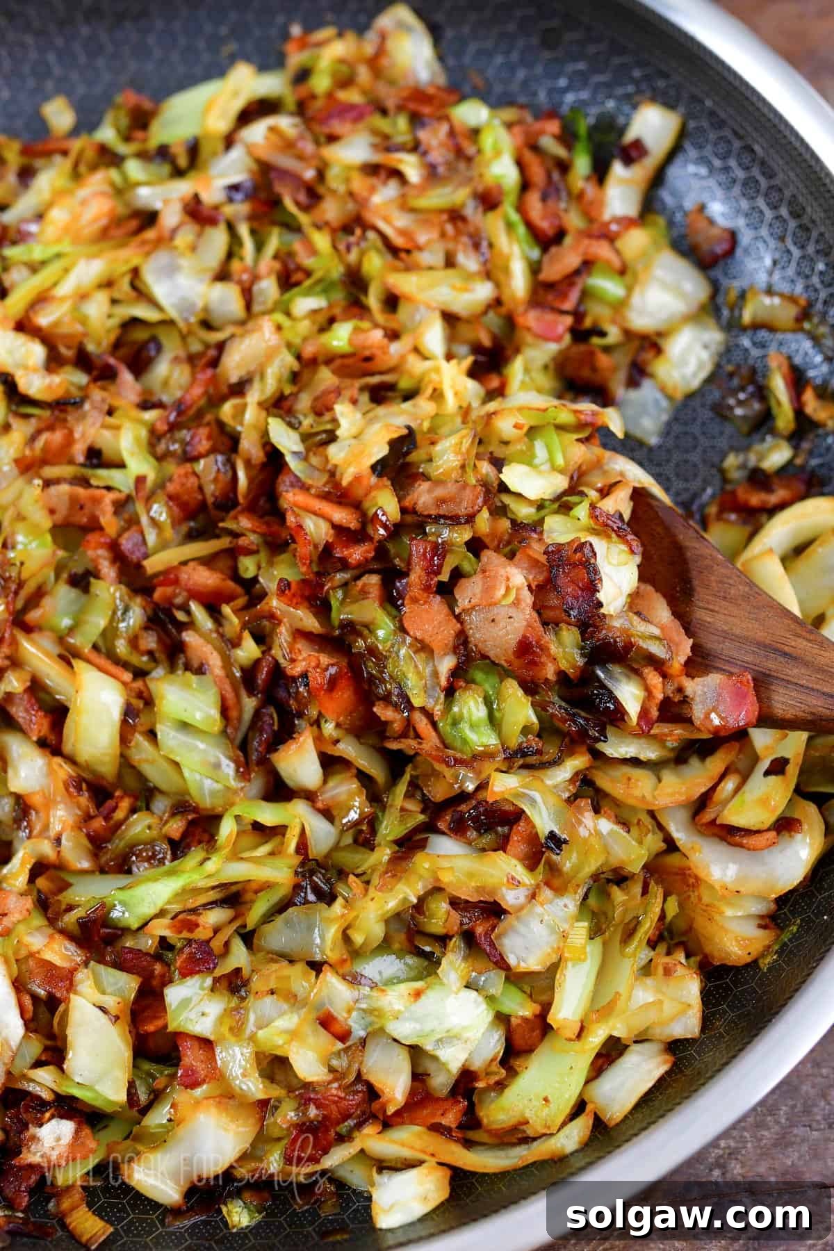 A serving spoon scoops golden-brown fried cabbage with crispy bacon and tender leeks from a large, hot skillet.