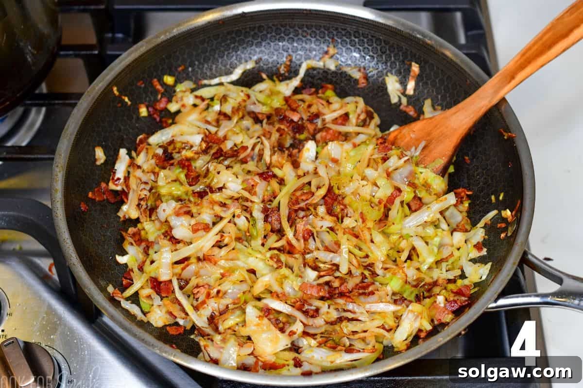 Close-up of golden-brown fried cabbage, leeks, and crispy bacon sizzling in a large skillet, stirred with a wooden spoon.