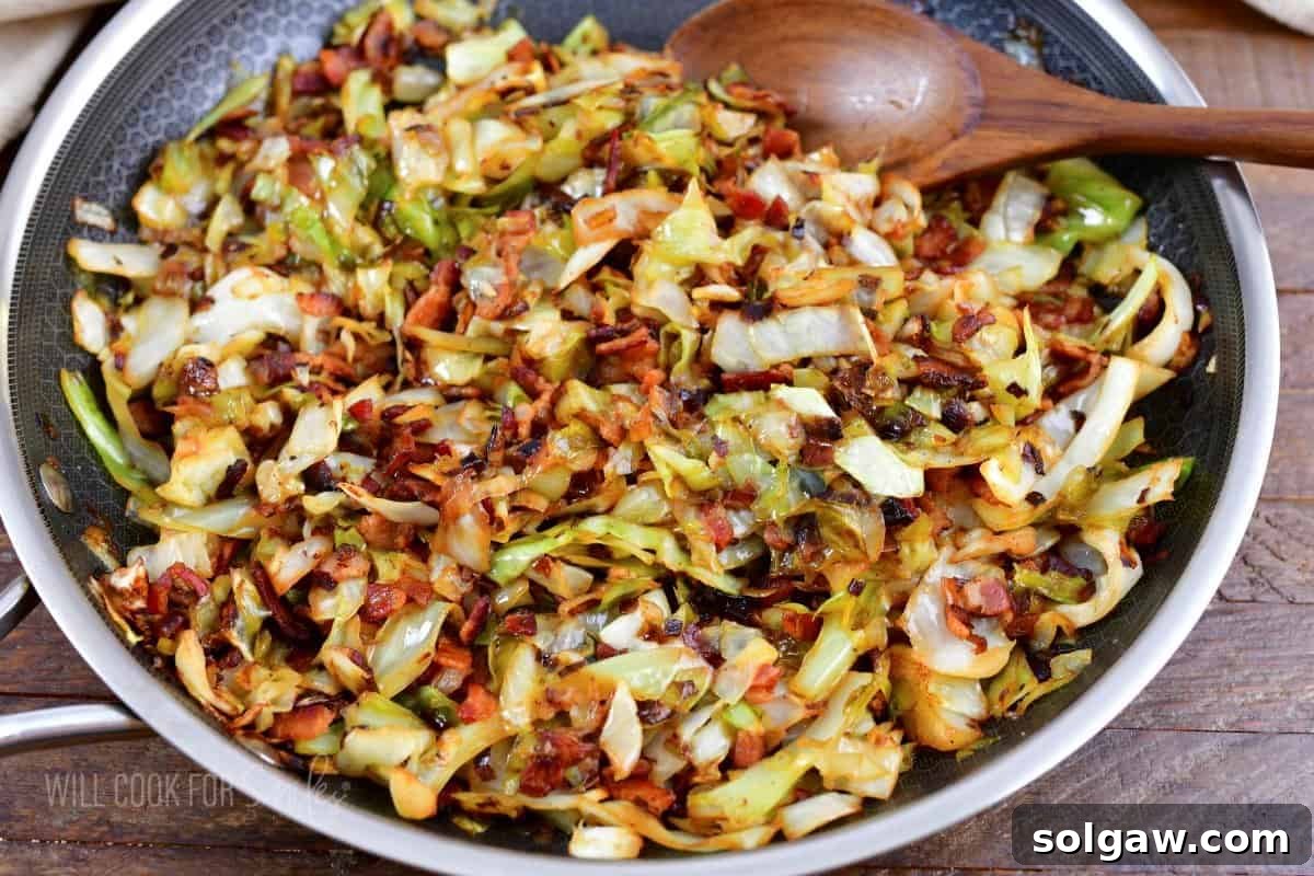 A close-up view of perfectly fried cabbage and crispy bacon in a cooking pan, with a spoon resting in it, ready to serve.