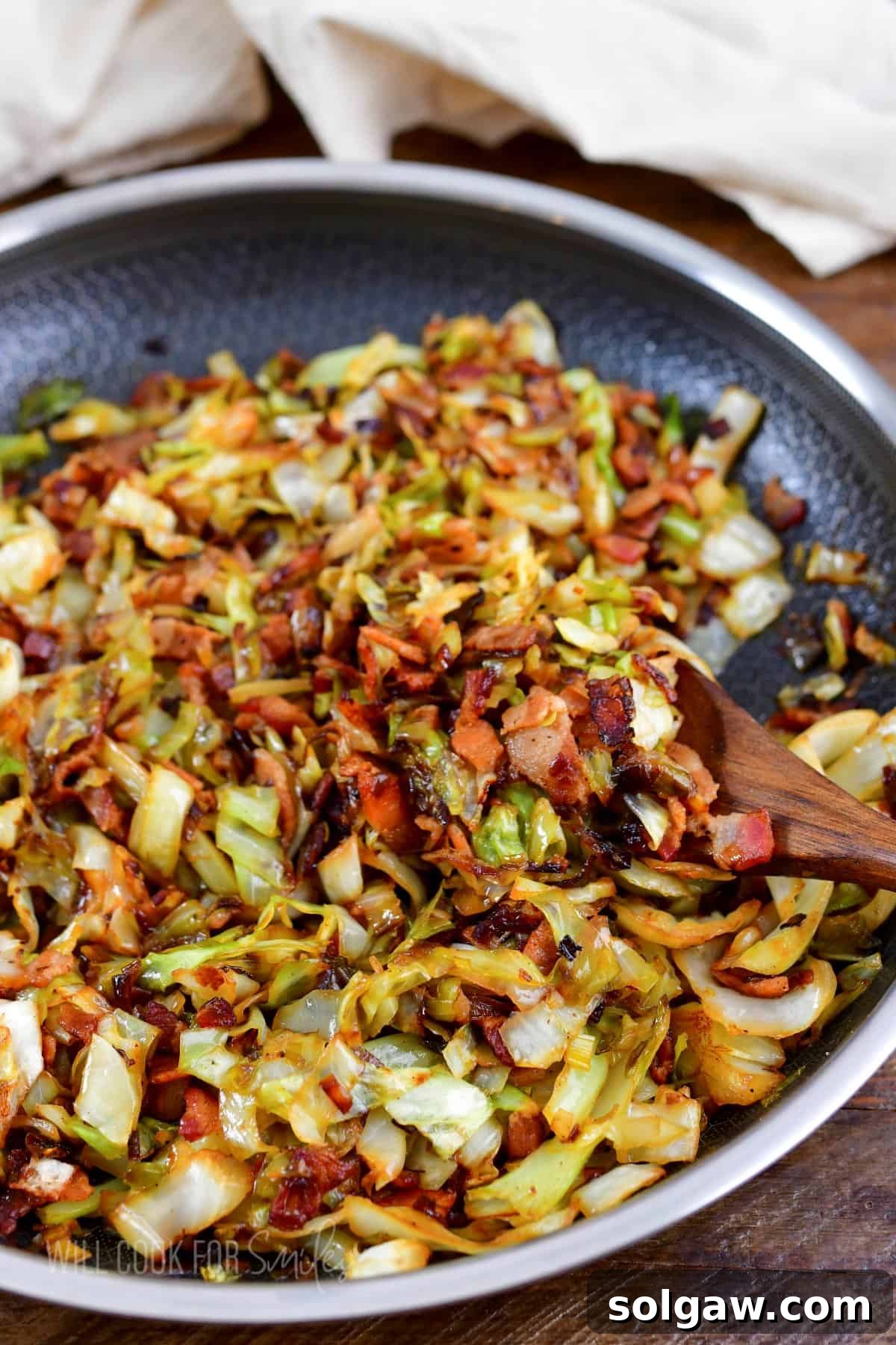 A wooden spoon lifts a generous serving of perfectly fried cabbage and crispy bacon from a large cooking pan.