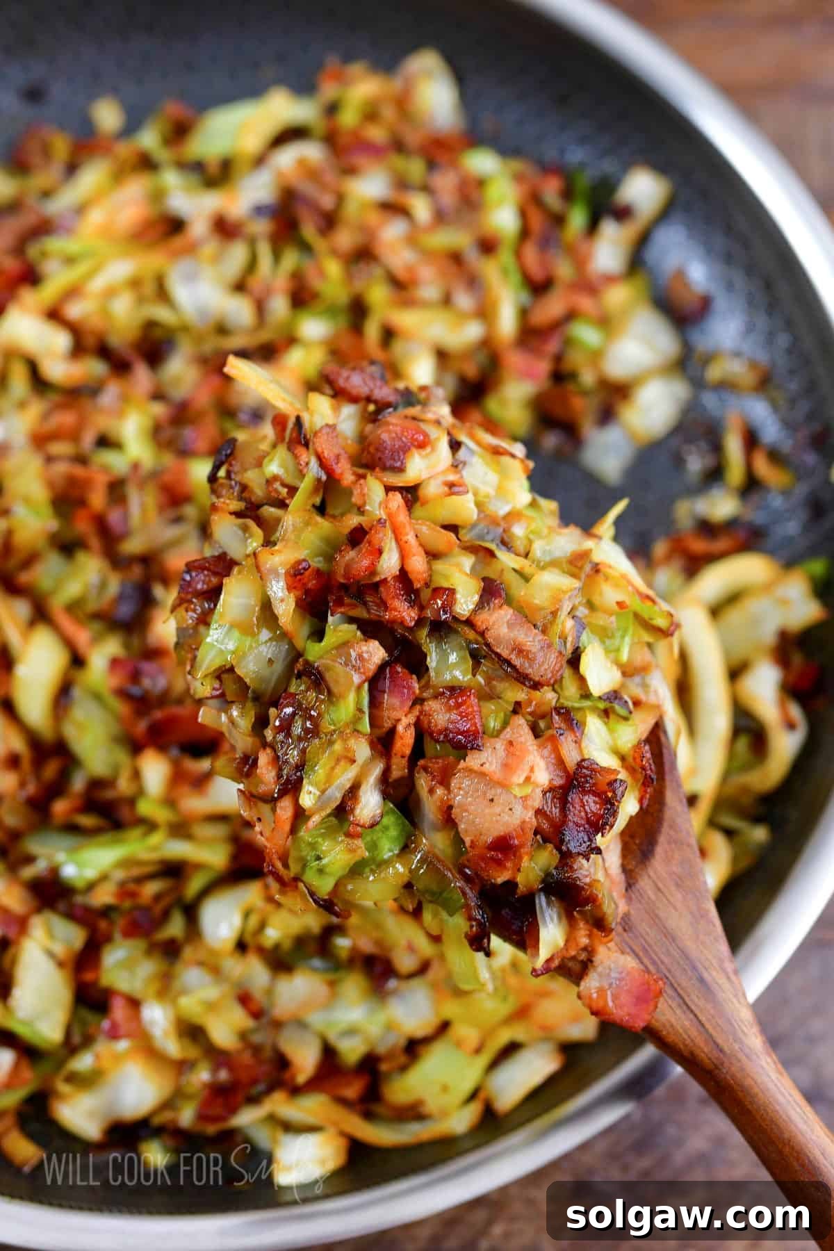 A close-up of a wooden spoon holding a perfect scoop of fried cabbage with crispy bacon and tender leeks, hovered above the cooking pan.