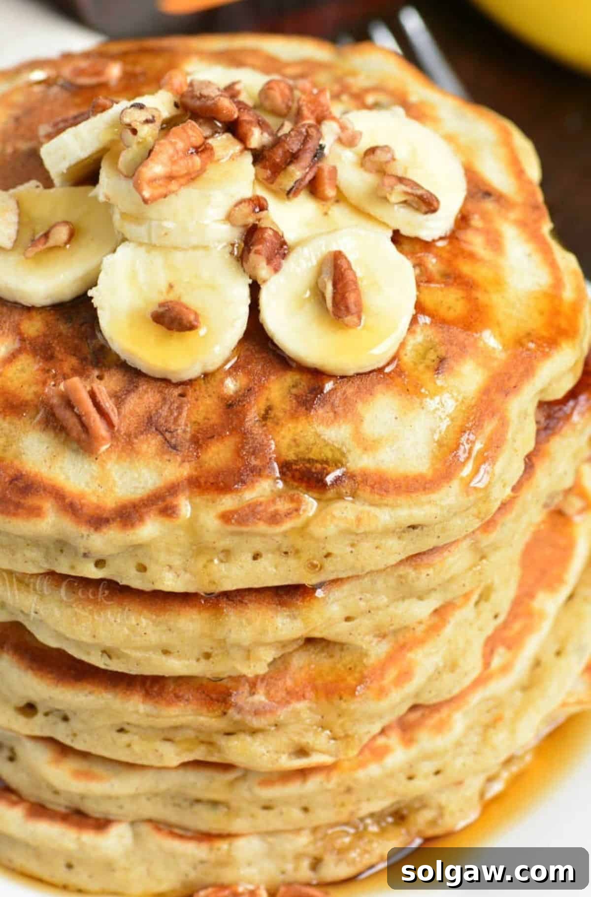 Stack of golden banana pancakes adorned with fresh banana slices, crunchy pecans, and a generous drizzle of maple syrup, ready to be enjoyed.