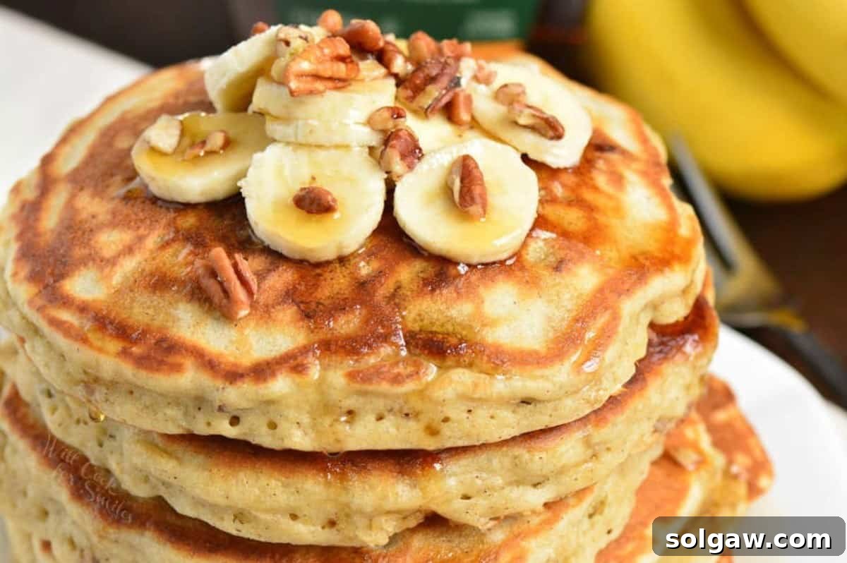 A close-up view of banana pancakes generously topped with thin banana slices, crunchy pecans, and glistening maple syrup, illustrating a delicious serving.