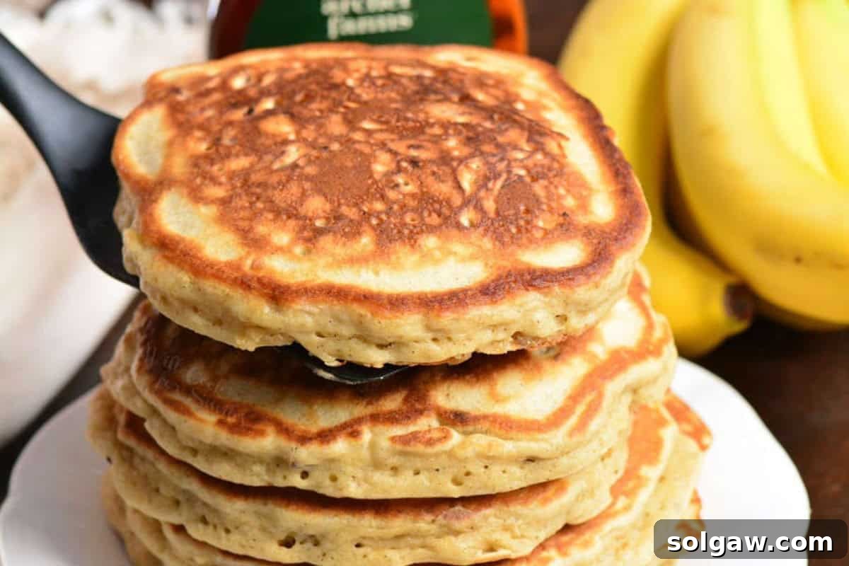 A hand adding another perfectly cooked banana pancake to a growing stack, with a lightly greased skillet in the background.