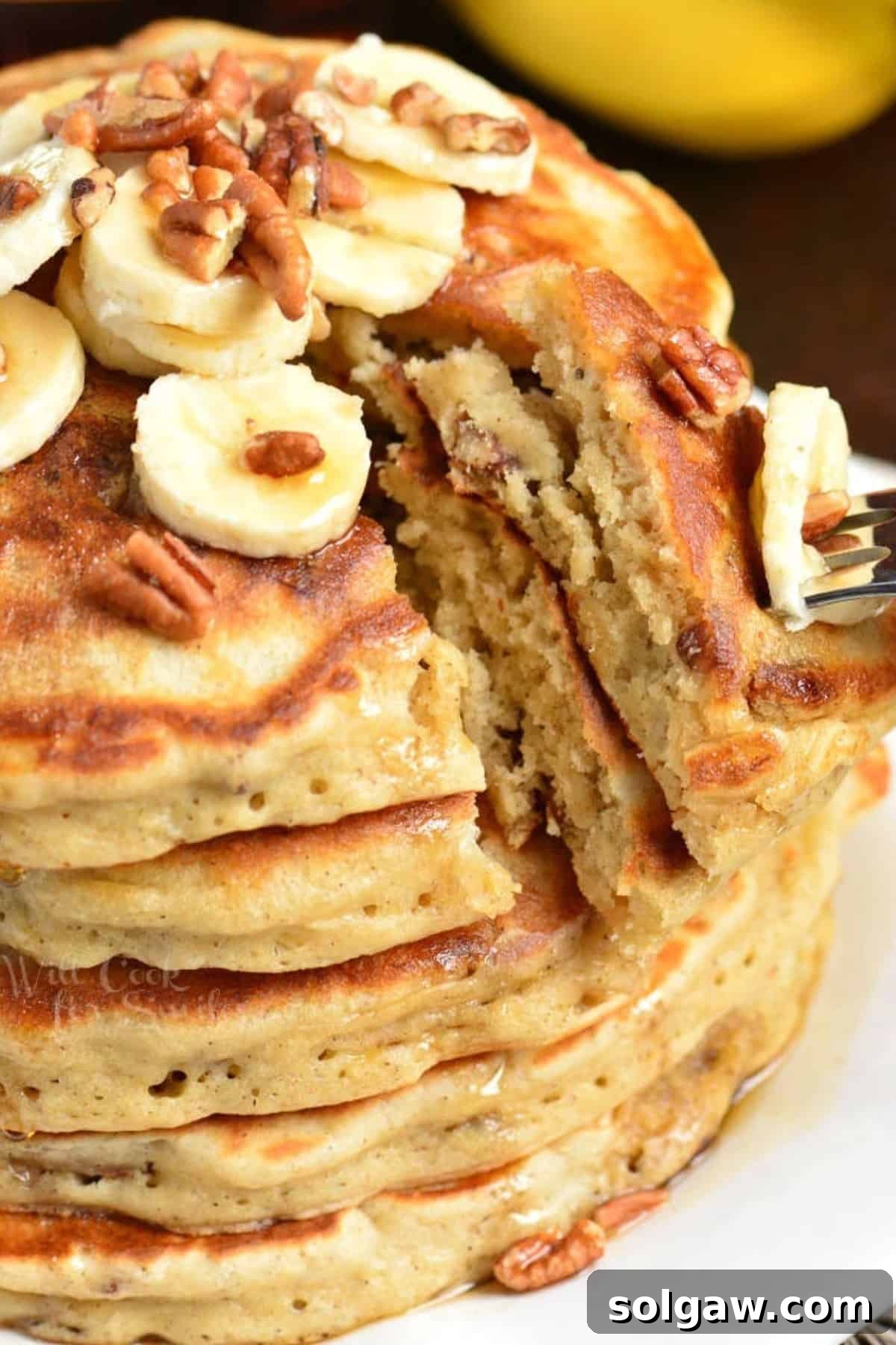 A fork cuts into a stack of banana pancakes, lifting a fluffy slice topped with nuts and syrup, ready for a bite.