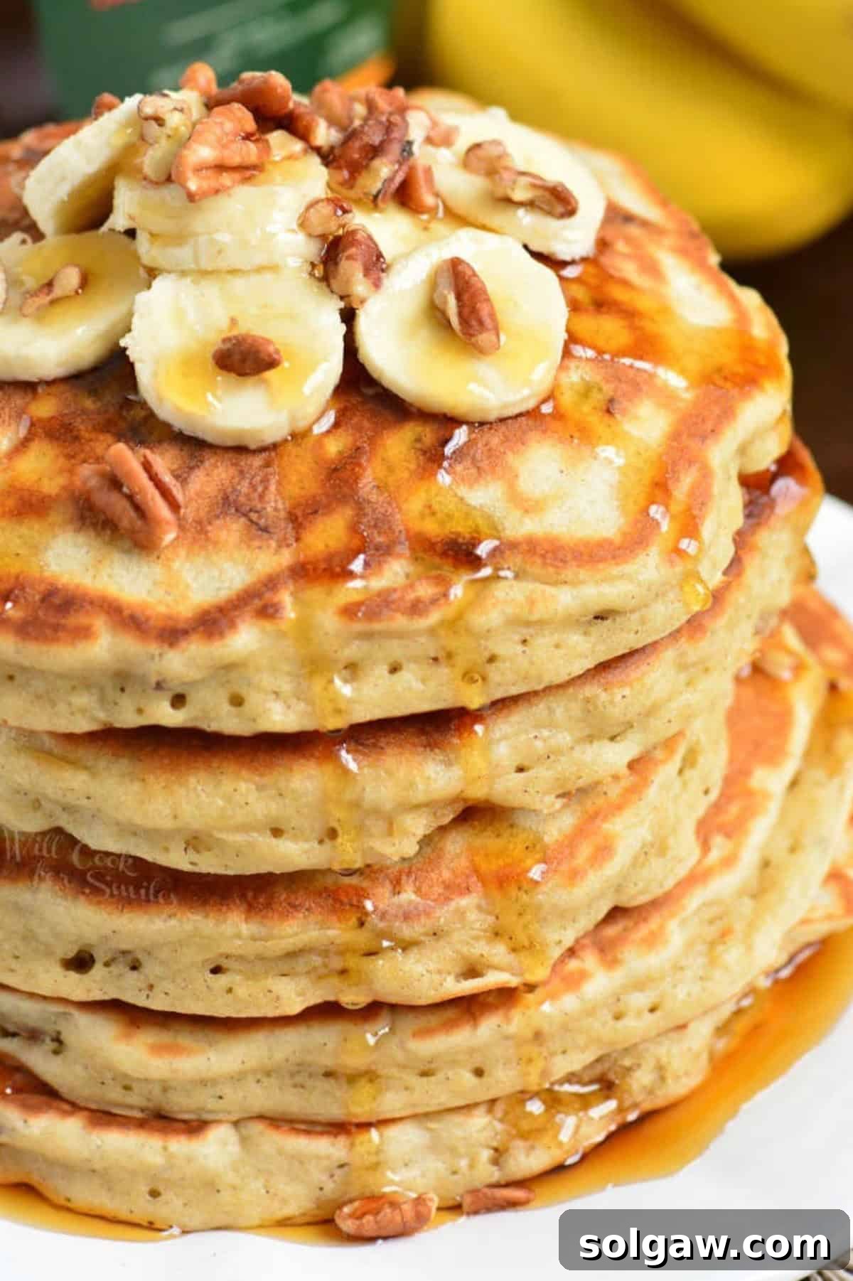 Stack of five golden banana pancakes beautifully arranged and topped with fresh banana slices, various nuts, and a generous pour of maple syrup, inviting a delightful breakfast.