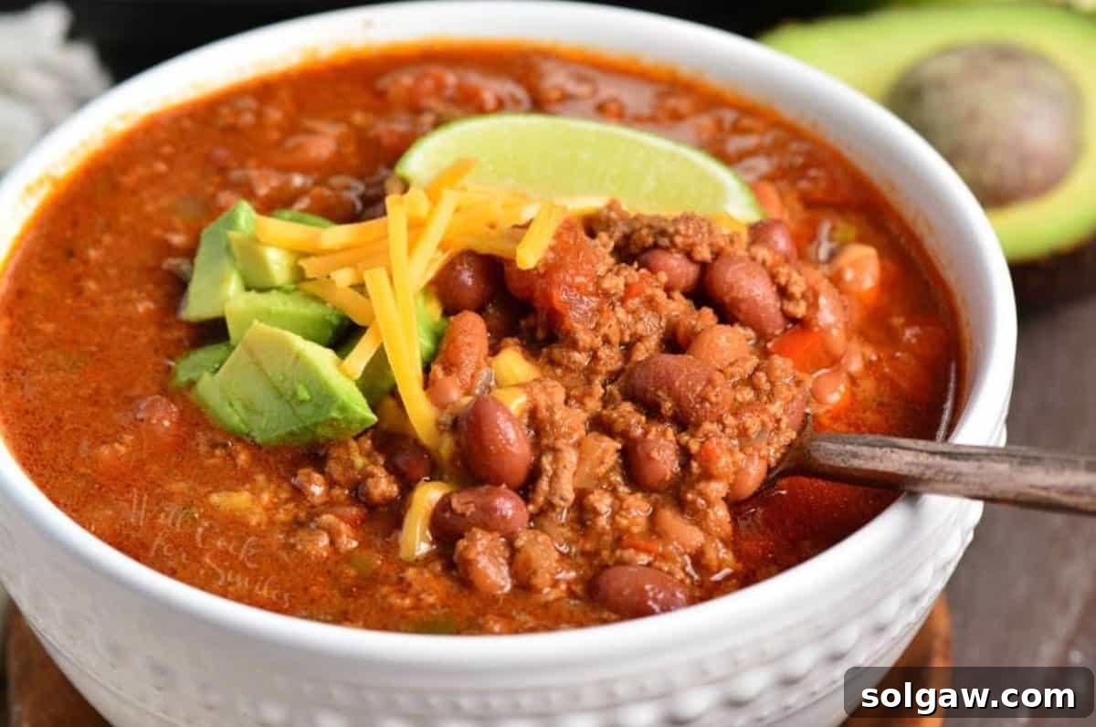 A perfectly portioned bowl of Instant Pot Chili, adorned with a dollop of sour cream, a sprinkle of shredded cheese, fresh diced avocado, and a vibrant lime wedge for garnish, with a spoon nestled in.