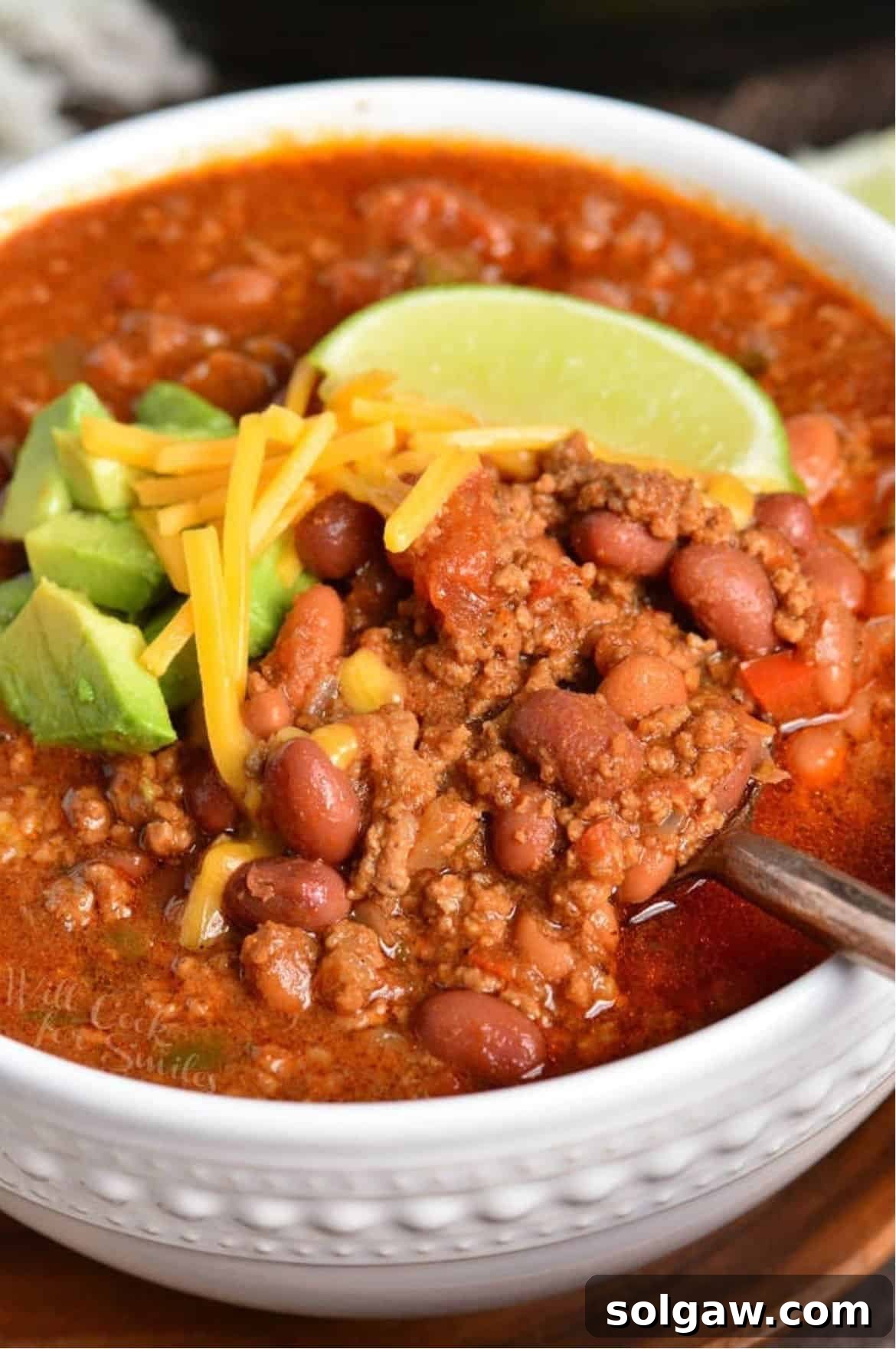 A close-up shot of Instant Pot chili in a white bowl, with a spoonful being lifted, showcasing the rich texture and garnishes of avocado, shredded cheese, and a lime wedge.