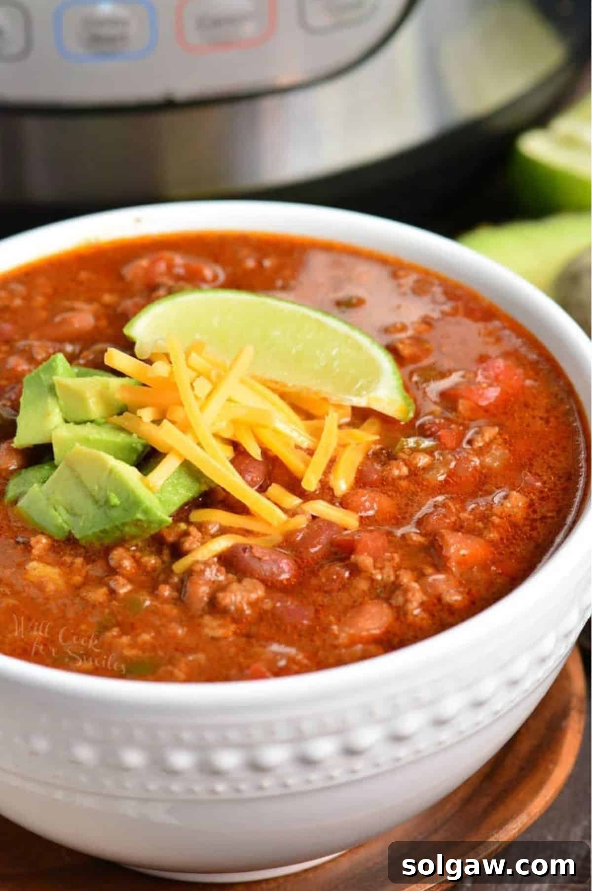 A beautifully presented bowl of Instant Pot Chili, served on a rustic wooden plate, topped generously with shredded cheese, fresh diced avocado, and a vibrant lime wedge.