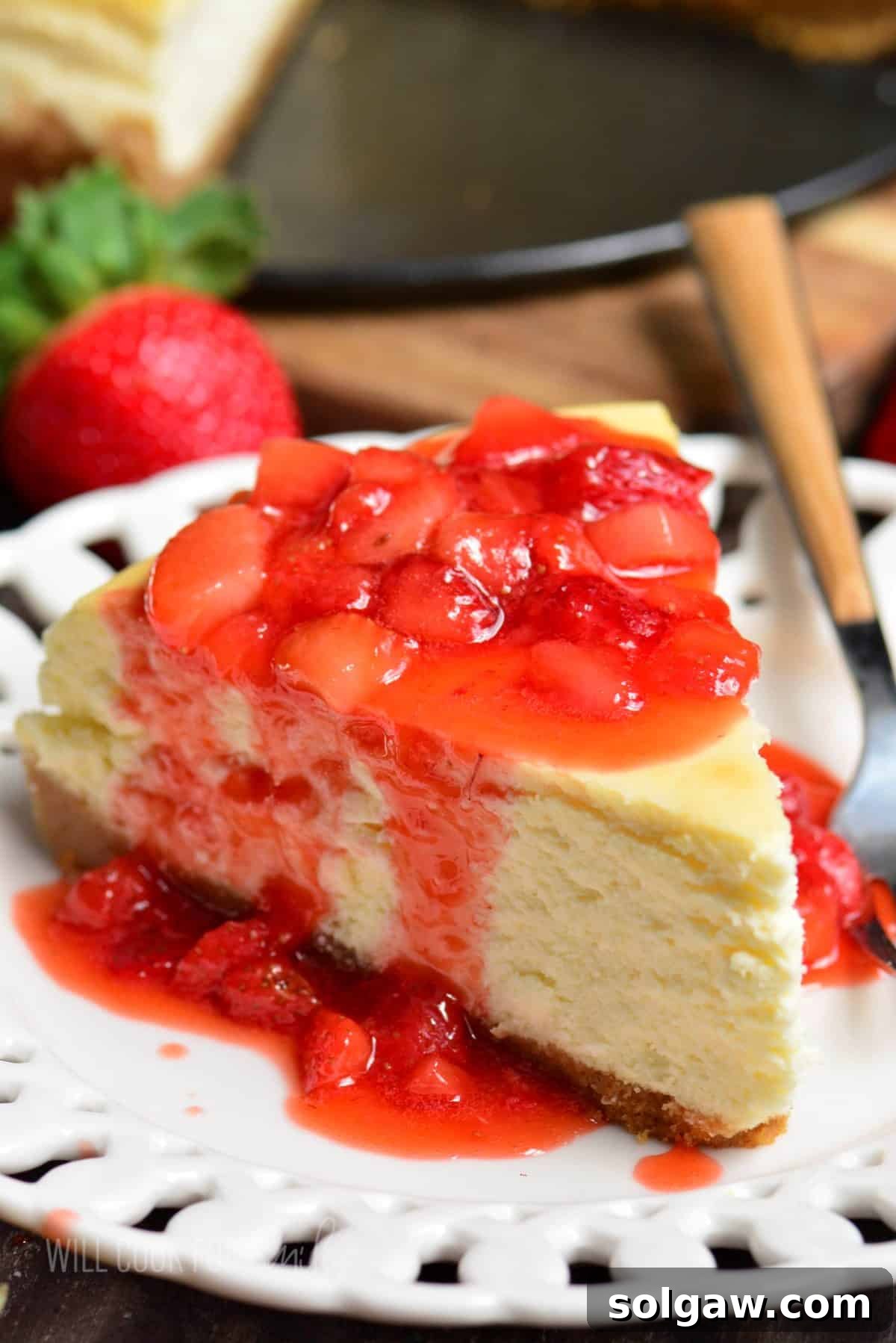 A beautiful slice of classic New York cheesecake generously topped with vibrant strawberry sauce on a white plate, highlighting its rich, creamy texture.