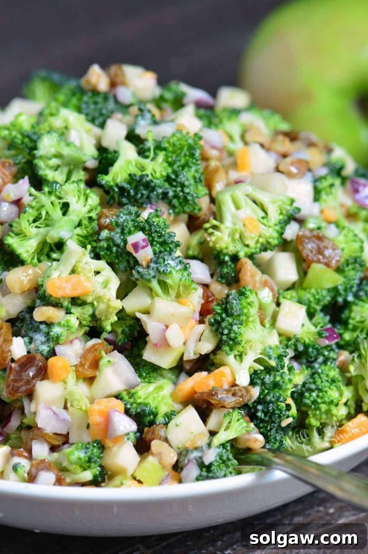 A vibrant tossed broccoli salad, rich with crisp green broccoli, sweet red apple chunks, golden raisins, and crunchy walnuts, is beautifully presented in a clean white bowl, ready to be served. 