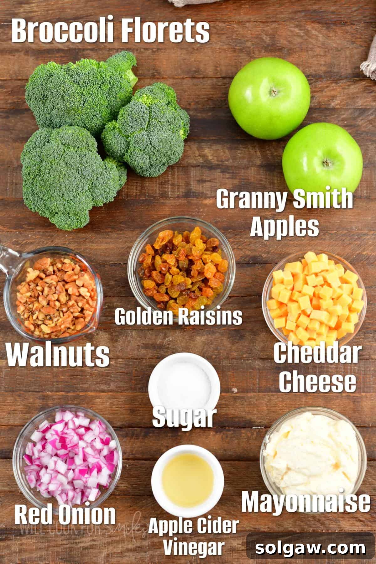 All the fresh ingredients for a delicious broccoli salad are neatly arranged on a rustic wooden surface, showcasing vibrant green broccoli, crisp red apples, golden raisins, walnuts, and red onion. 
