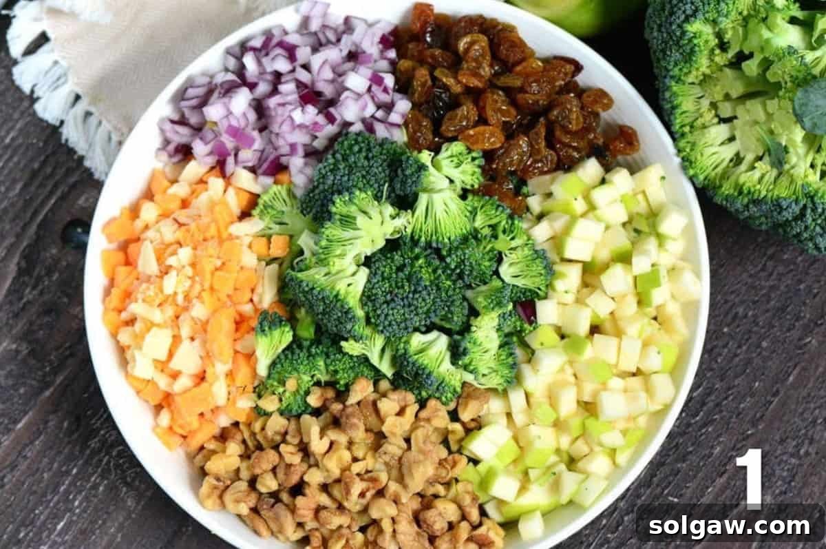 All the prepared ingredients for apple broccoli salad, including chopped broccoli, diced apples, raisins, walnuts, red onion, and cheddar cheese, are combined in a large bowl, ready for the dressing.