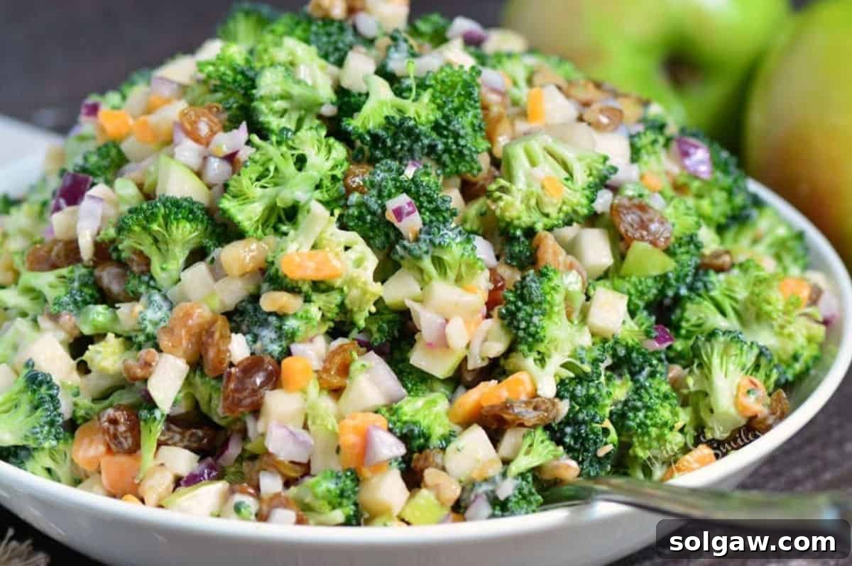 A vibrant and refreshing apple broccoli salad, generously mixed with a creamy dressing, is elegantly presented with a large serving spoon in a pristine white bowl. 