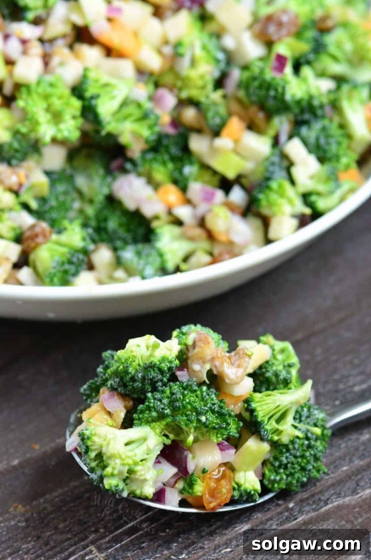 A serving spoon filled with a generous scoop of apple broccoli salad, showcasing crisp broccoli, diced apples, and a creamy dressing, is ready to be enjoyed from a white bowl.