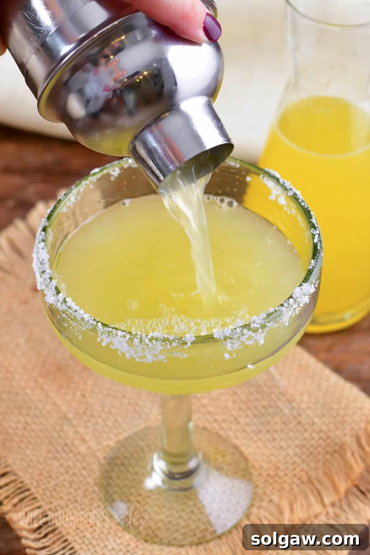 A freshly made margarita being poured from a cocktail shaker into a salt-rimmed glass.