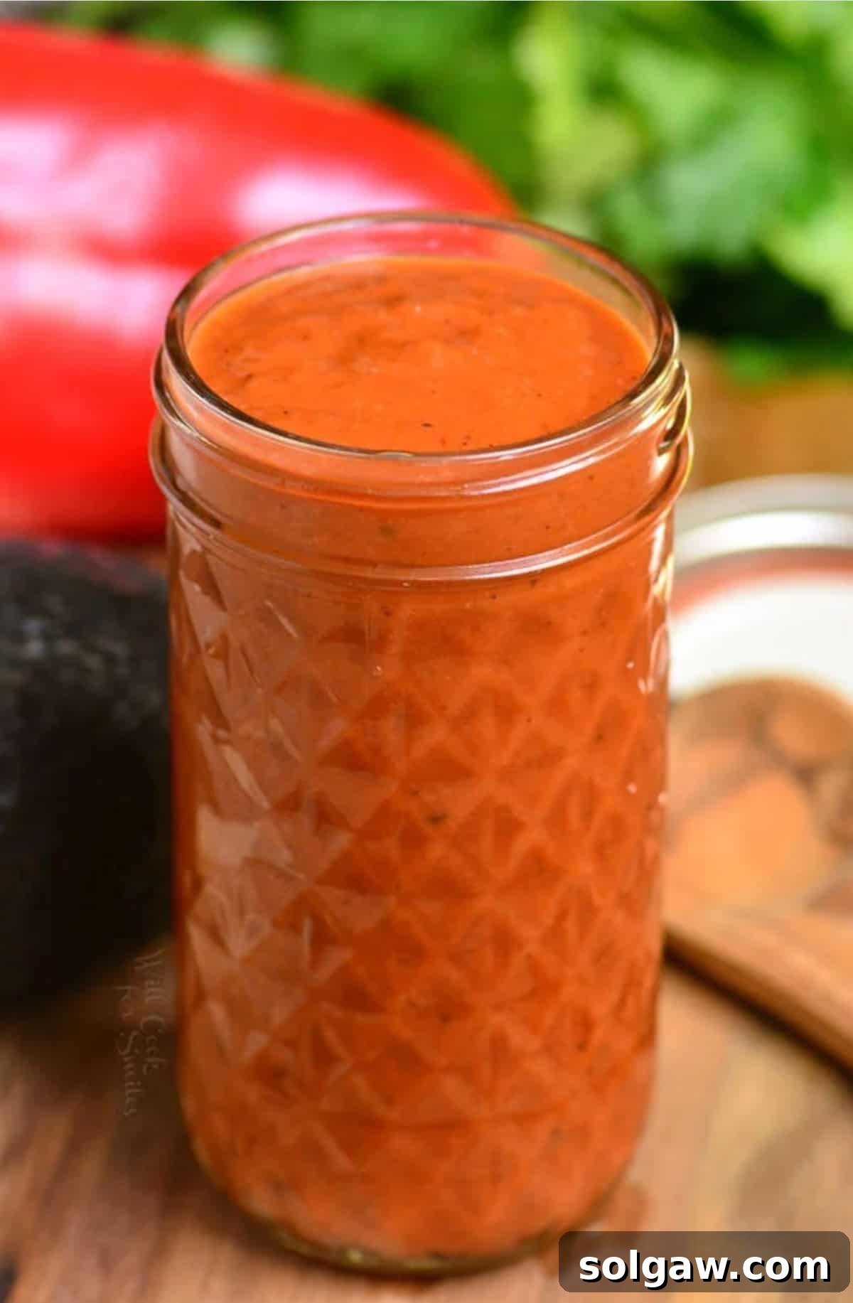 Homemade Enchilada Sauce 2 Tall glass jar full of rich homemade enchilada sauce on a wooden surface with fresh vegetables like tomatoes, onions, and chili peppers arranged around it, highlighting the vibrant color and fresh ingredients.