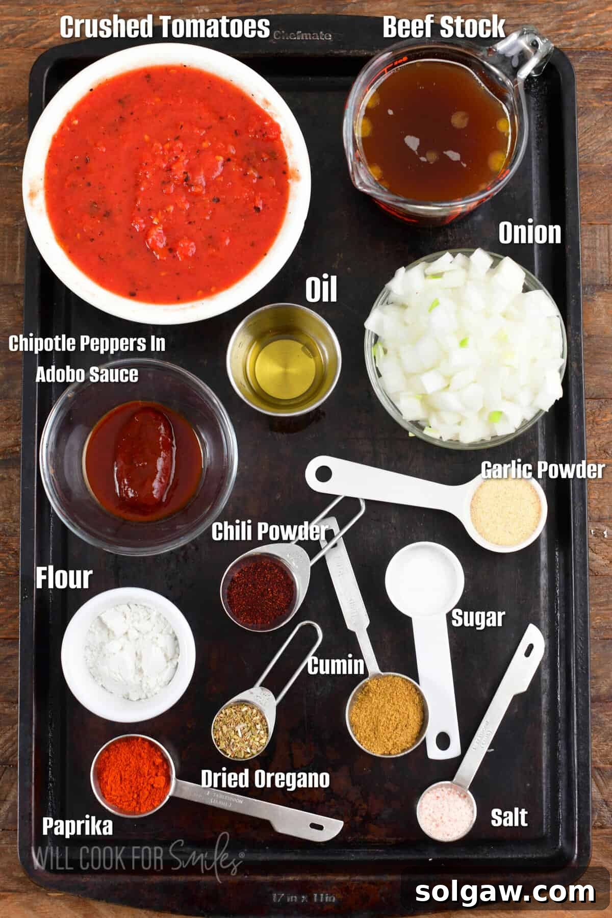 Homemade Enchilada Sauce 3 A collection of fresh and dried ingredients laid out on a metal sheet tray, ready to make homemade enchilada sauce, including a yellow onion, a can of crushed tomatoes, a jar of chipotles in adobo, and various spices like chili powder, cumin, and oregano.