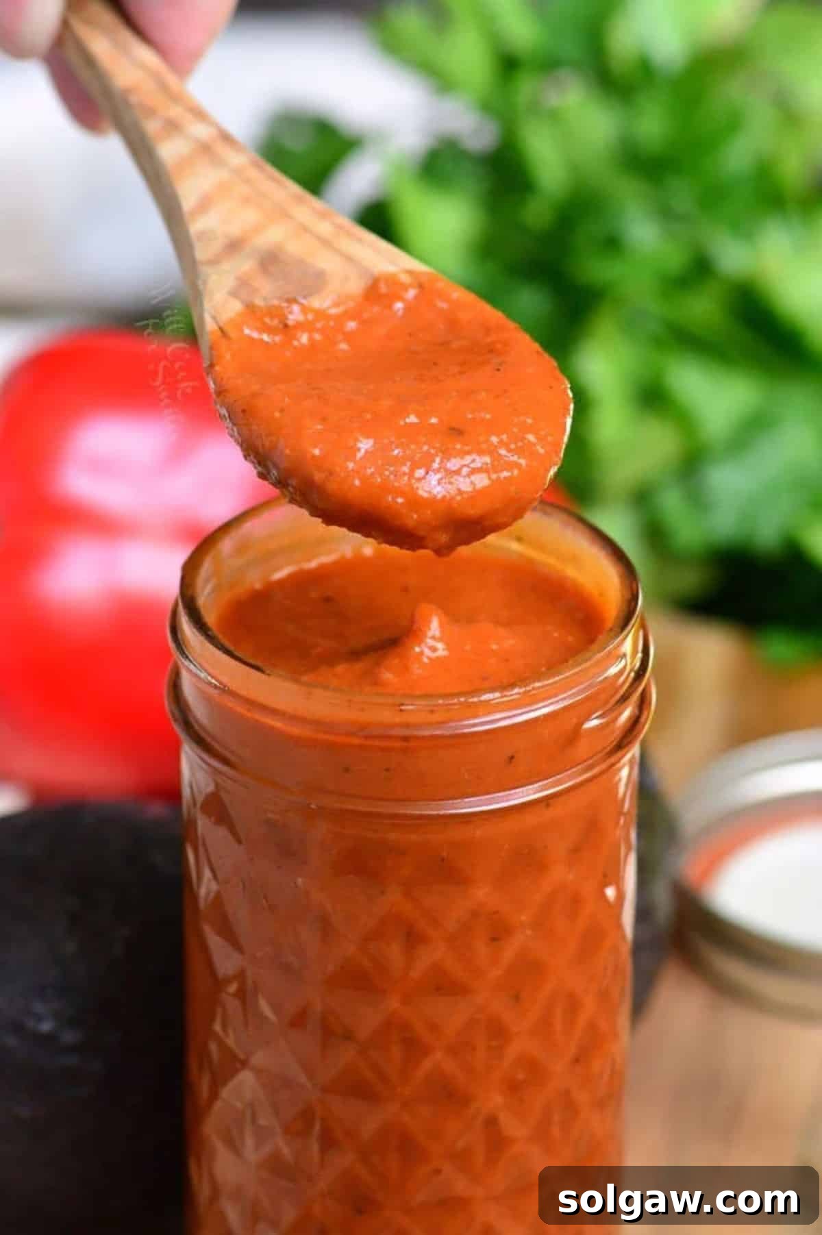 Homemade Enchilada Sauce 6 A wooden spoon lifting a spoonful of thick, homemade enchilada sauce directly over a clear glass jar filled with the vibrant red sauce, illustrating its smooth texture and rich consistency.