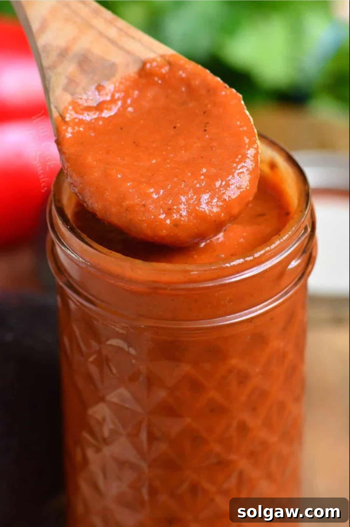 Homemade Enchilada Sauce 8 A close-up of a wooden spoon scooping savory homemade enchilada sauce from a glass jar, emphasizing its smooth texture and rich, deep red color, ready for use in any dish.