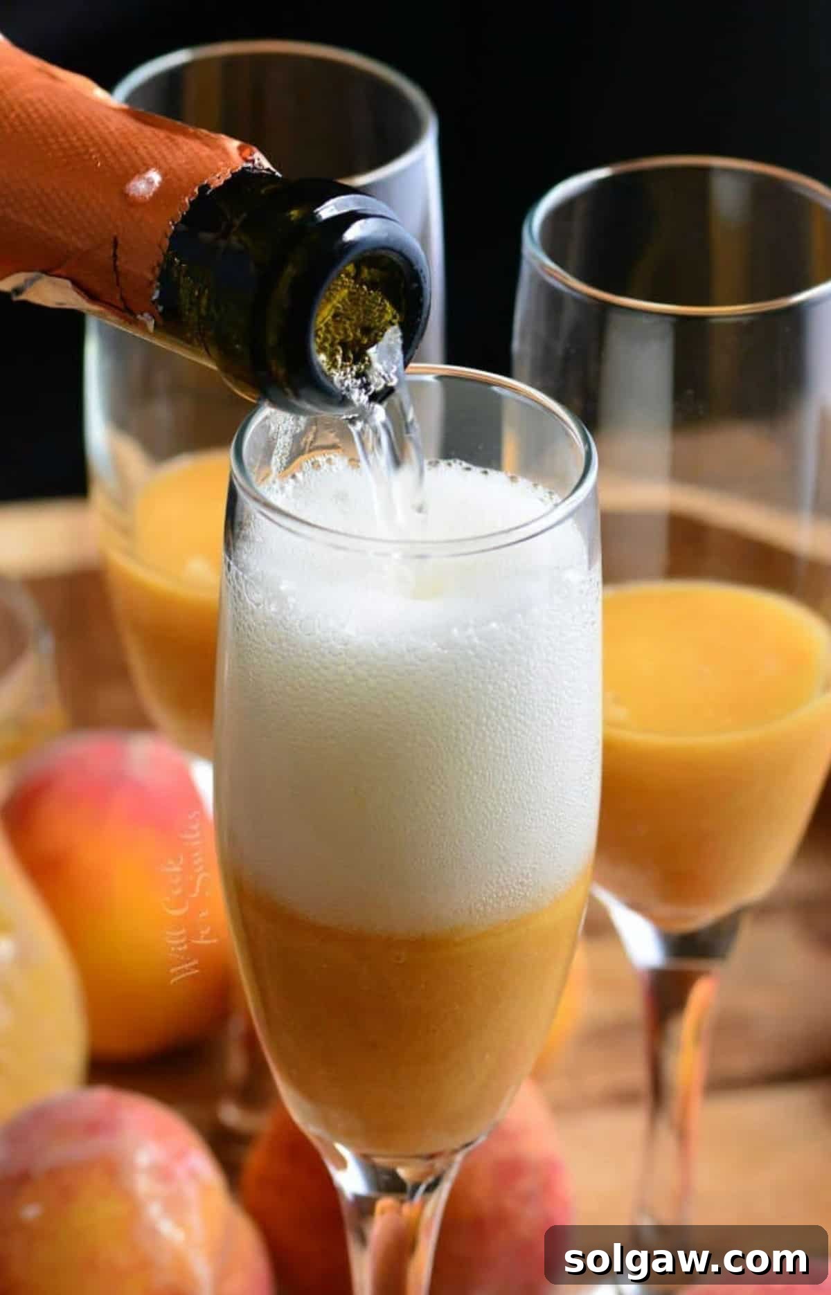 A steady hand pours Prosecco into a champagne flute already containing fresh peach puree, demonstrating the angled pouring technique to minimize foam.